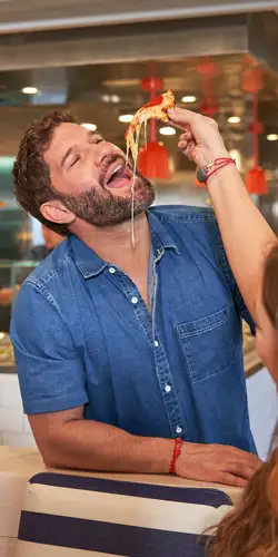 A husky white guy with a beard being fed pizza at the Virgin Voyages pool